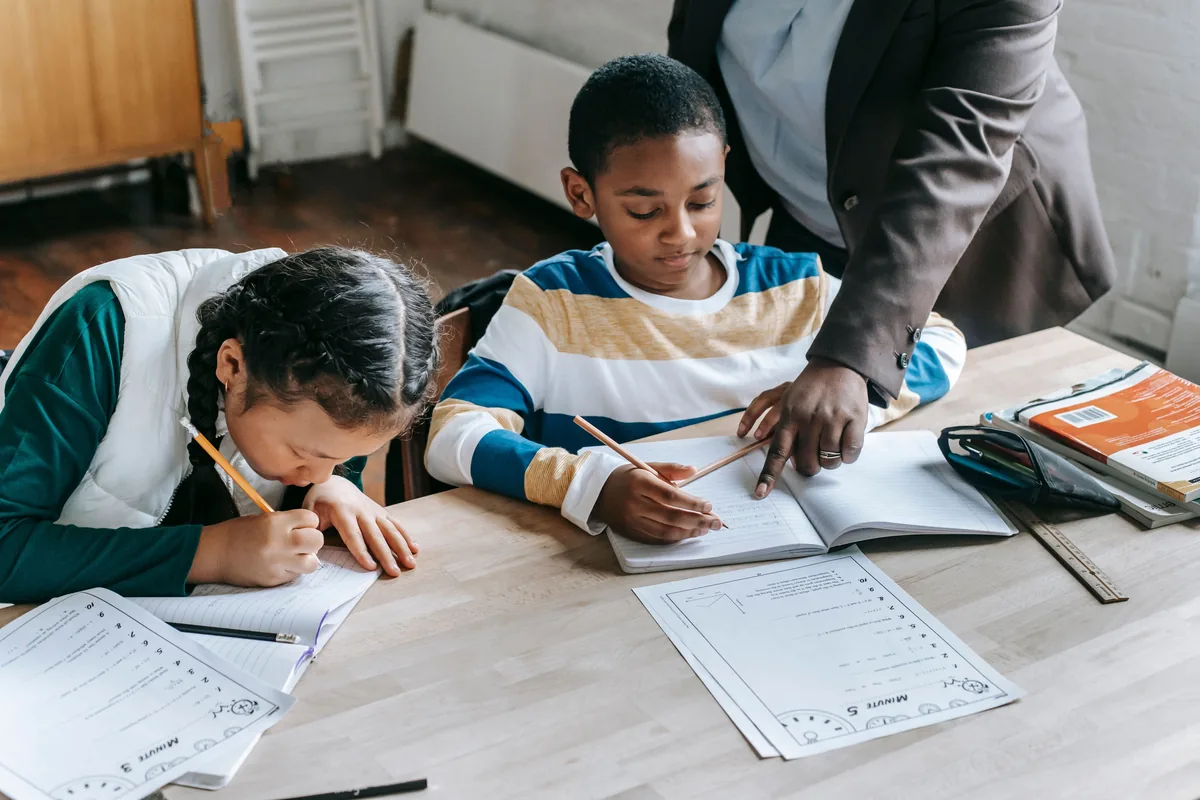 A teacher points to a workbook while two students work attentively